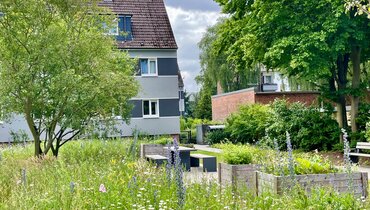 Blütenreiche Grünfläche mit Hochbeeten und Sitzgelegenheiten vor einem Wohngebäude. Die naturnah gestaltete Außenanlage bietet Lebensraum für Insekten und Aufenthaltsmöglichkeiten für Anwohnende.